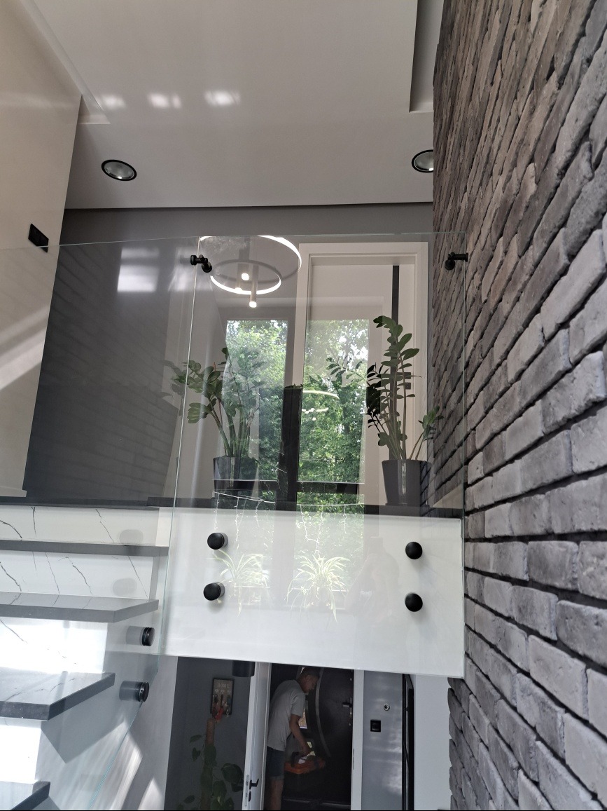 Szklana balustrada schodowa z czarnymi mocowaniami, widok z dołu. Fragment ściany z cegły, rośliny doniczkowe i schody z marmurowym wzorem.