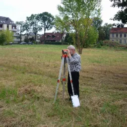 Geodeta w koszuli w kratę, z teodolitem na łące, mierzy teren pod budowę. W tle budynki mieszkalne i drzewa.
