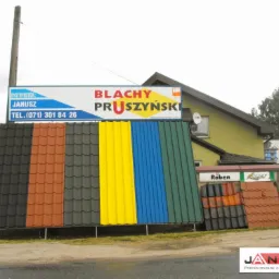 Ekspozycja dach&oacute;wek i blach Pruszyński w r&oacute;żnych kolorach, z widocznym szyldem dystrybutora Janusz i fragmentami budynk&oacute;w w tle. Prezentacja materiał&oacute;w.