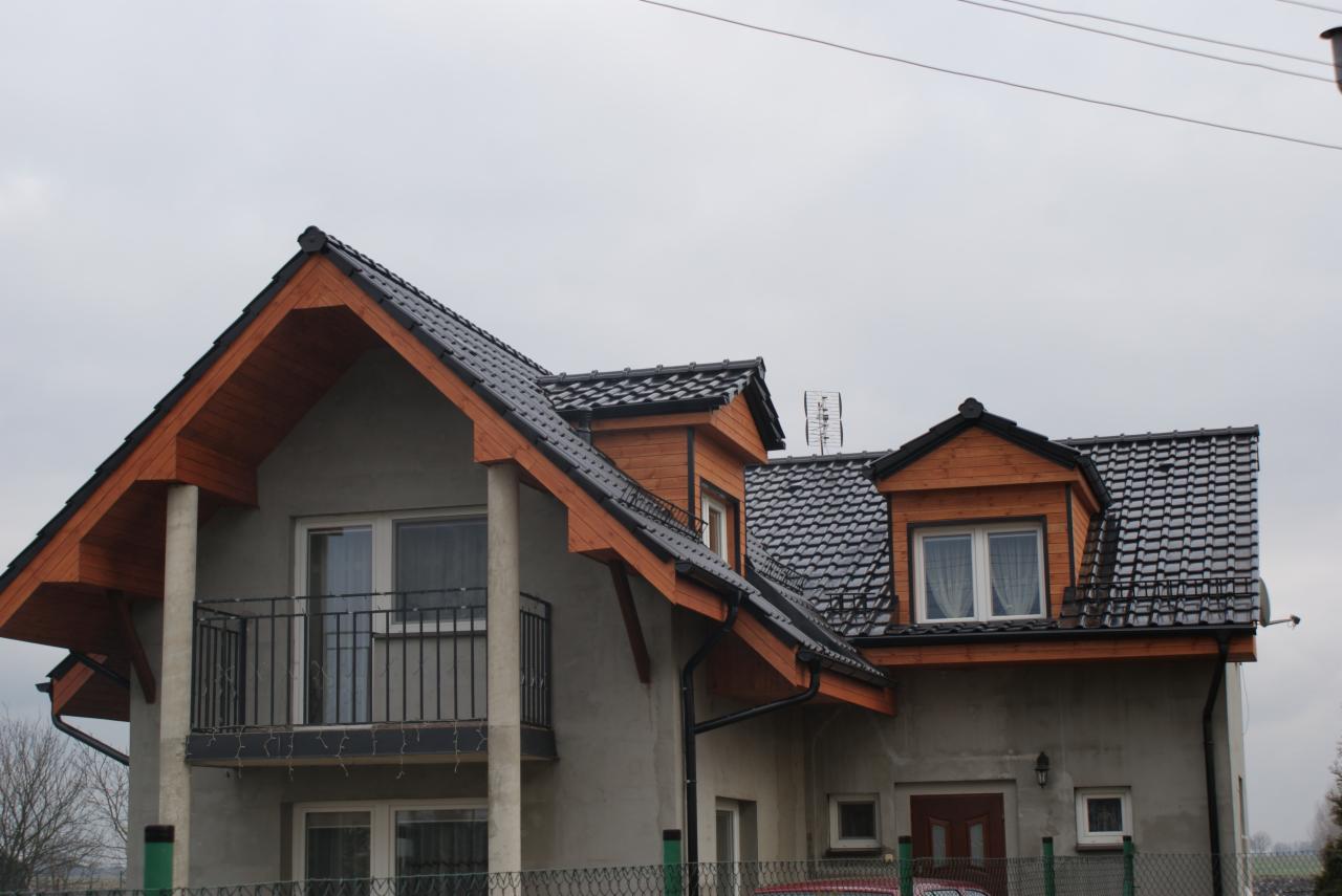 Dom jednorodzinny z ciemną dachówką i drewnianymi elementami wykończeniowymi. Widoczne okna dachowe i balkon z metalową balustradą. Pochmurne niebo w tle.