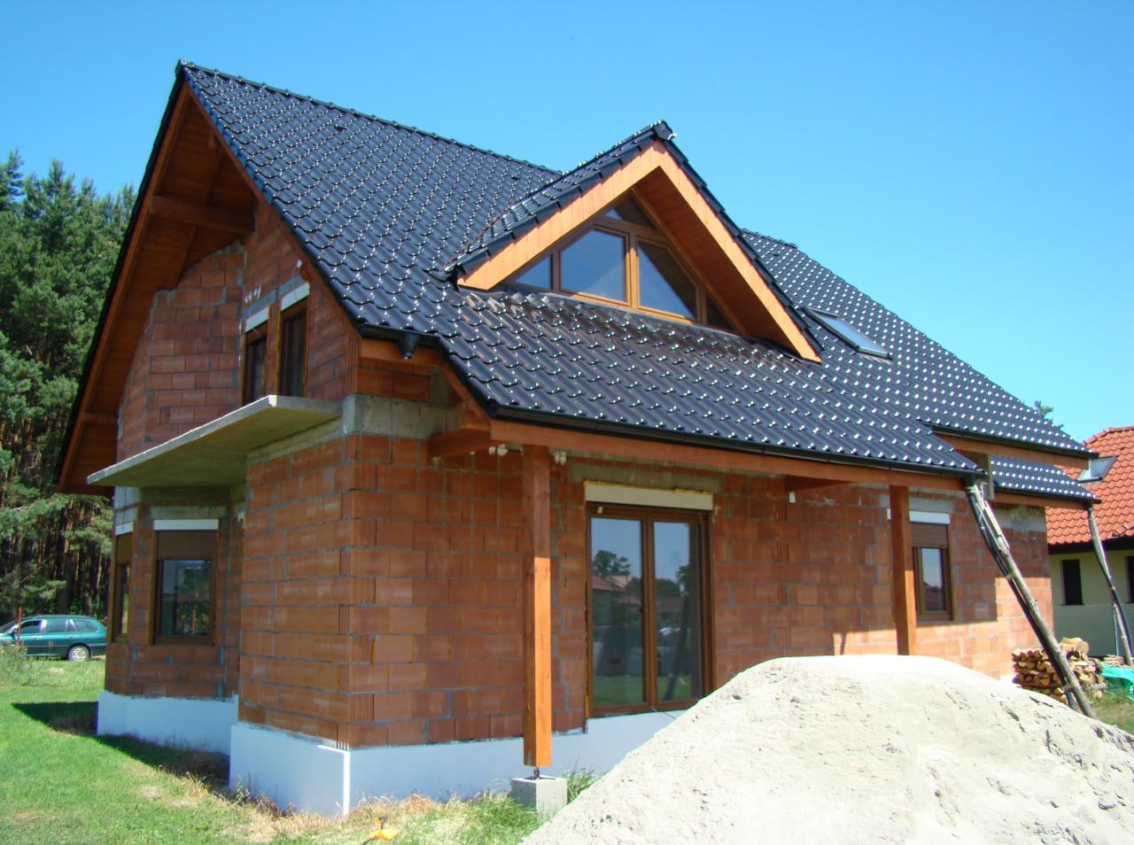 Dom w trakcie budowy z czarnym dachem z dachówki ceramicznej, widoczne cegły i drewniane elementy konstrukcyjne, na tle błękitnego nieba i zieleni.