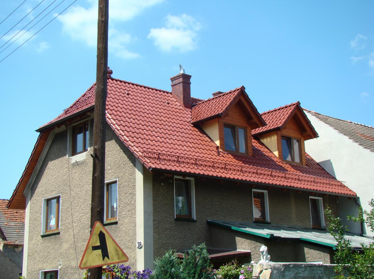 Dom z nową, czerwoną dachówką ceramiczną i drewnianymi lukarnami, widziany z poziomu ulicy. Na pierwszym planie znak drogowy i słup energetyczny.