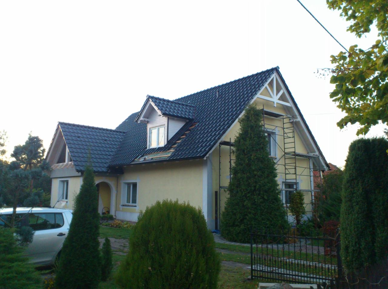 Dom z żółtą elewacją i ciemną dachówką, częściowo osłonięty rusztowaniem. Fragment dachu w trakcie prac, widoczne elementy konstrukcyjne.  Zieleń w otoczeniu.
