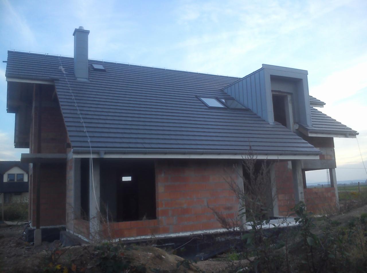 Szary dach z dachówką ceramiczną i oknami połaciowymi na nowym domu w Oławie. Kominek z blachy. Budynek w stanie surowym otwartym.
