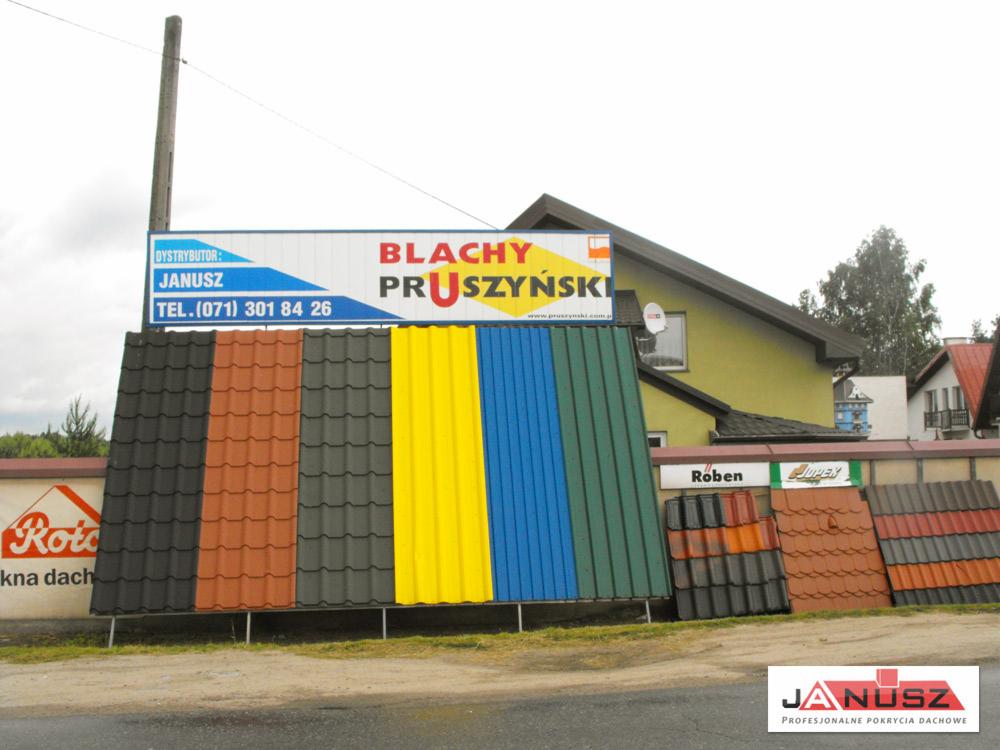 Ekspozycja dachówek i blach Pruszyński w różnych kolorach, z widocznym szyldem dystrybutora Janusz i fragmentami budynków w tle. Prezentacja materiałów.