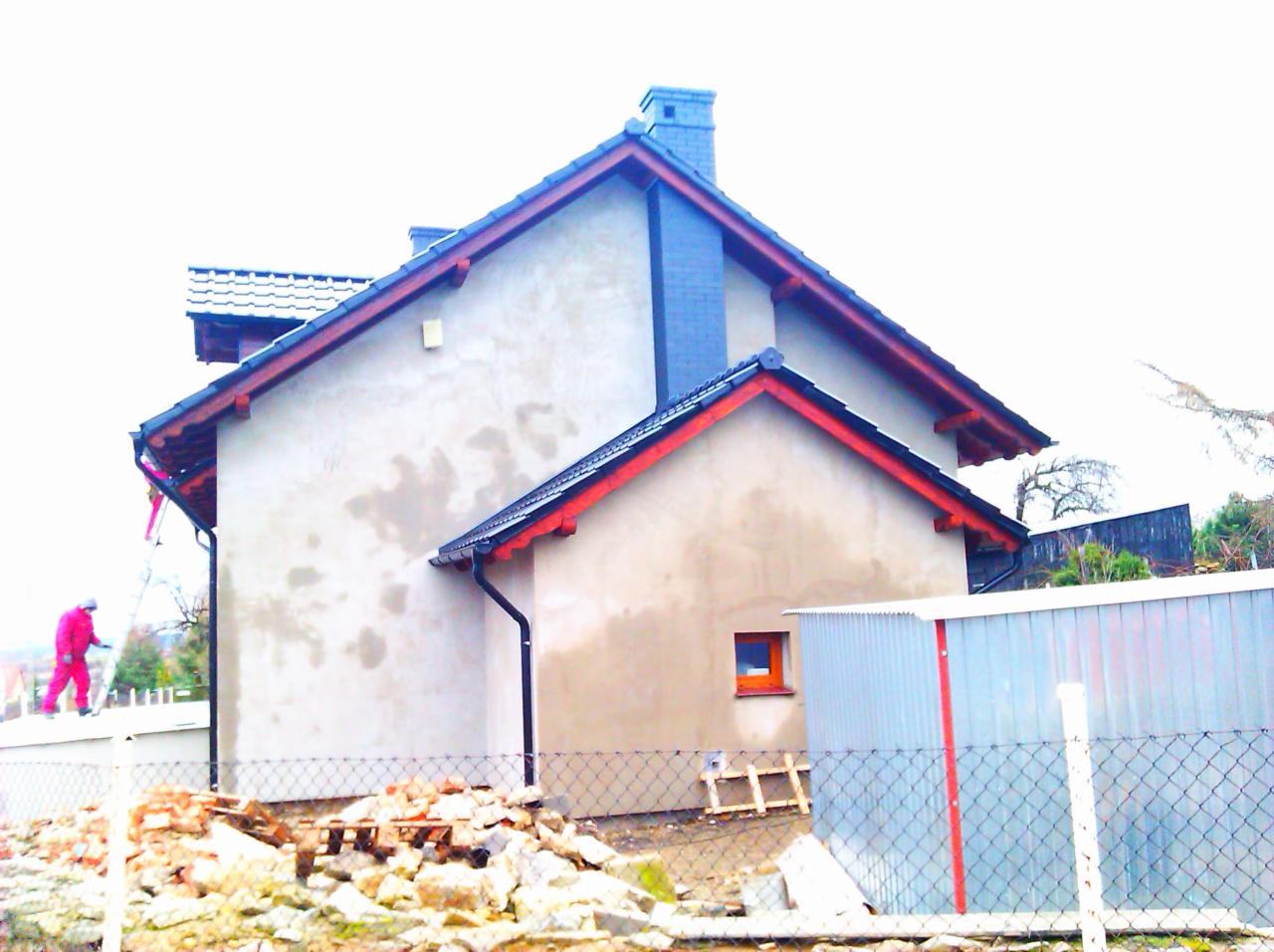 Dwukondygnacyjny dom z szarą elewacją i czarnym dachem. Czerwone wykończenia dachu. Na dachu pracownik w czerwonym kombinezonie. Na pierwszym planie gruz i fragment ogrodzenia.