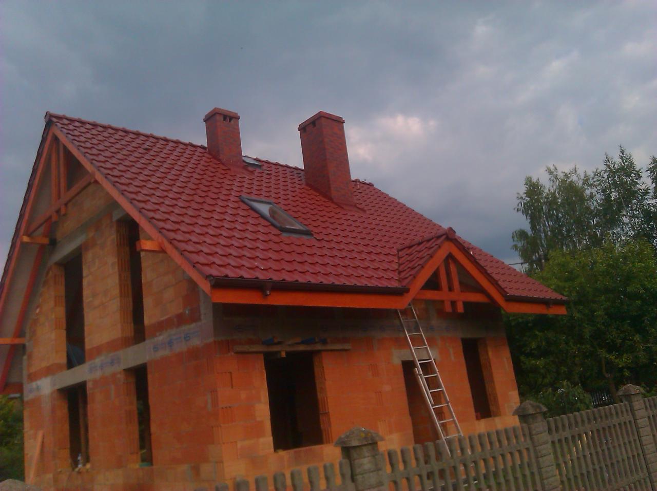 Dom w trakcie budowy z czerwonym dachem z dachówki, widoczne kominy, okno dachowe i drabina oparta o ścianę. Pochmurne niebo w tle.