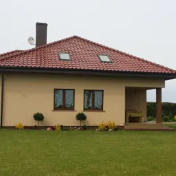 Parterowy dom jednorodzinny z czerwoną dachówką, dwoma oknami dachowymi i beżową elewacją, widok z zadbanego trawnika z niskimi krzewami ozdobnymi.