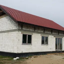 Niedokończony dom jednorodzinny z czerwonym dachem, widoczne białe bloczki konstrukcyjne, okna i drzwi balkonowe wstawione, teren wok&oacute;ł niezagospodarowany, w tle drugi dom w budowie.