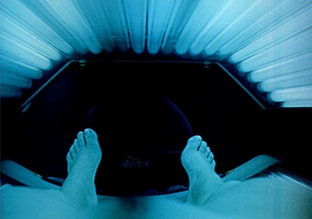 Widok z perspektywy leżącej osoby w solarium, widoczne stopy i lampy emitujące niebieskie światło.