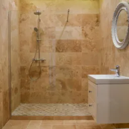 Wykończona łazienka z kabiną prysznicową typu walk-in, beżowe płytki trawertynowe na ścianach i podłodze, mozaika w strefie prysznicowej, biała szafka z umywalką i okrągłe lustro w ozdobnej ramie.