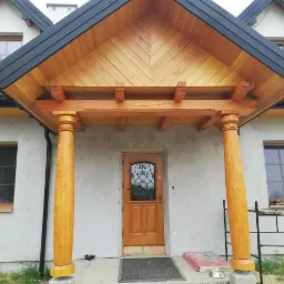 Drewniany ganek z kolumnami przed wejściem do domu z szarą elewacją i czarnym dachem.