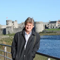 Mężczyzna w kurtce na tle zamku nad rzeką, w słoneczny dzień. Elementy architektury w tle, zieleń i woda. Uśmiechnięty mężczyzna oparty o barierkę.