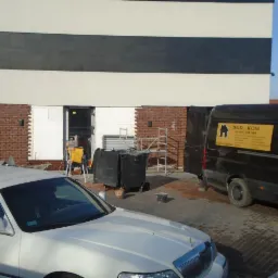 Budowa domu: fragment elewacji z cegły klinkierowej w trakcie montażu, widoczny styropian izolacyjny, rusztowanie, samochód dostawczy Bud-Kom i biała limuzyna.