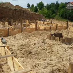 Szalunki fundamentów pod budowę domu jednorodzinnego w Kartuzach. Widoczne drewniane formy i zbrojenia w ziemi. Prace ziemne w toku, w tle zieleń i budynek.