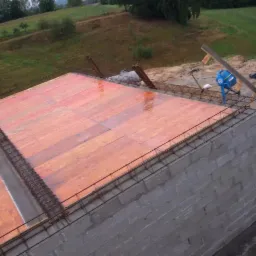 Wznoszenie ścian z bloczków betonowych z deskowaniem i zbrojeniem na górze, widok z góry. W tle łąka i drzewa. Mieszarka do betonu w tle.