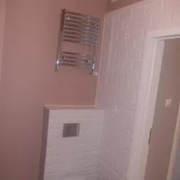 Wykończona łazienka z białą ceramiką i grzejnikiem drabinkowym na ścianie w kolorze pudrowego różu. Białe, prostokątne płytki imitujące cegłę.