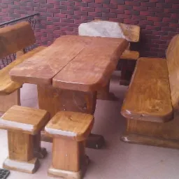 Rustykalny komplet mebli ogrodowych z surowego drewna: stół z dwoma blatami, ławy z oparciami i dwa stołki. Tło stanowi ceglana ściana i balustrada.