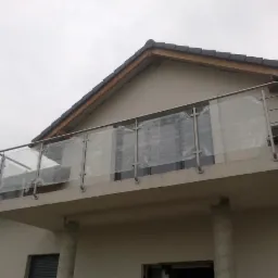 Szklana balustrada balkonowa ze stali nierdzewnej, zabezpieczona folią ochronną podczas montażu, widok z dołu na tle budynku z ciemną dachówką.