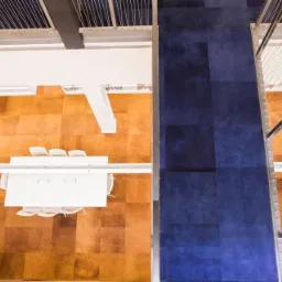 Widok z góry na dwa rodzaje podłóg: brązowe panele i niebieska wykładzina w biurze. Białe stoły i krzesła na panelach, metalowe barierki oddzielają podłogi.