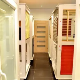 Wnętrze sklepu z ekspozycją okien i drzwi, widok na drewniane drzwi z przeszkleniami w głębi korytarza, z roletami w różnych kolorach po bokach.