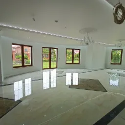 Przestronne wnętrze z marmurową posadzką w odcieniach beżu i brązu, z dekoracyjnymi wstawkami i refleksami światła odbijającymi okna. Białe ściany, ozdobne listwy sufitowe i kryształowe żyrandole...