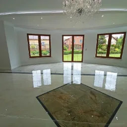 Salon z błyszczącą, beżowo-brązową podłogą z płytek, z dekoracyjnymi ciemnymi intarsjami, oświetleniem punktowym w suficie i kryształowym żyrandolem, z widokiem na ogród przez trzy okna...