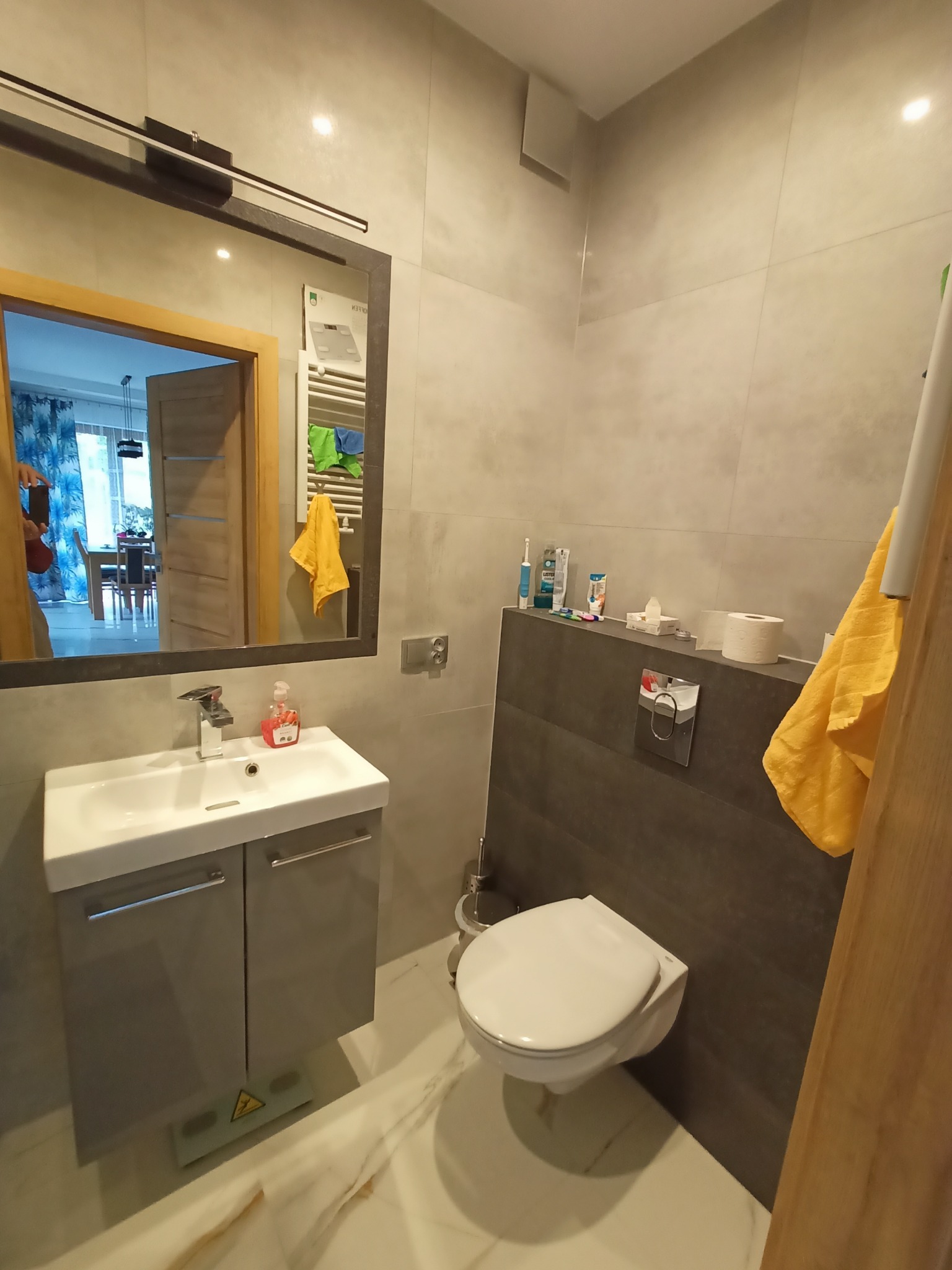 Szara łazienka z umywalką na szafce, podwieszaną toaletą i dużym lustrem, w którym odbija się widok na pokój z drewnianym stołem.