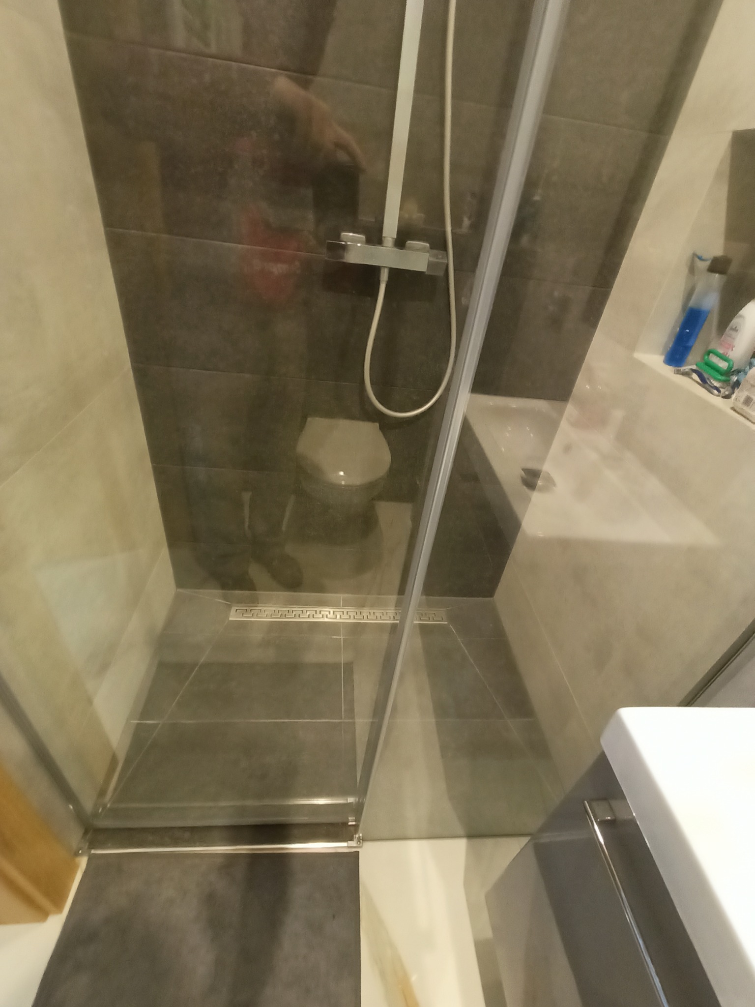 Nowoczesna kabina prysznicowa z odpływem liniowym, szare płytki na ścianach i podłodze, widoczne akcesoria łazienkowe na półce.