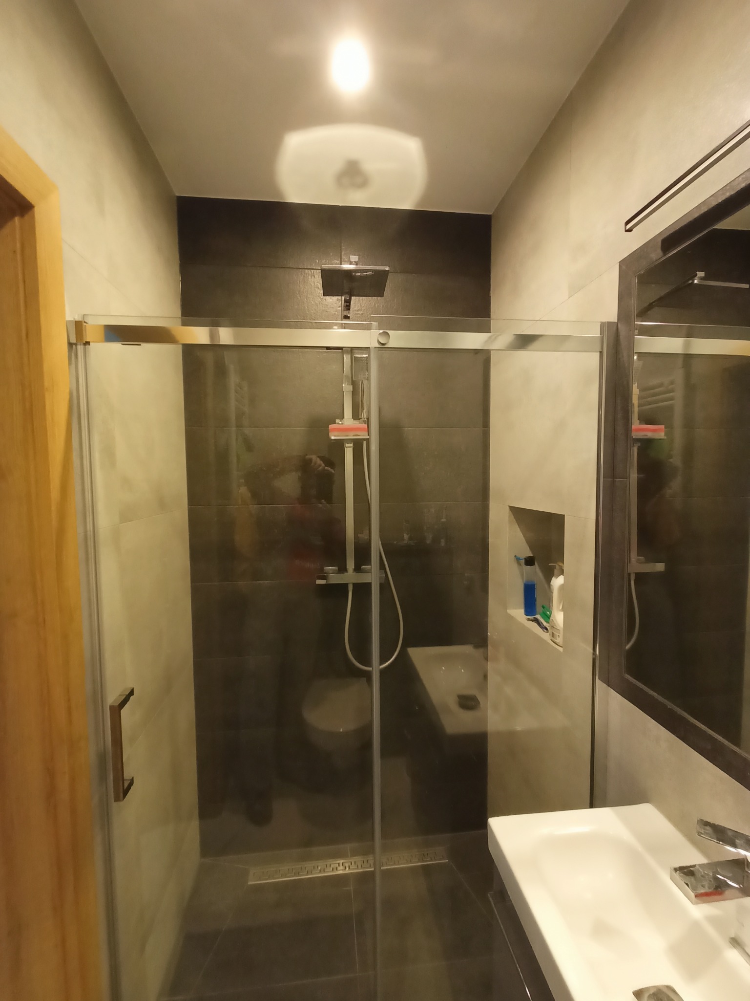 Nowoczesna łazienka z kabiną prysznicową typu walk-in, szare płytki, biała umywalka i wnęka z kosmetykami, widoczne odbicie fotografa w szkle.