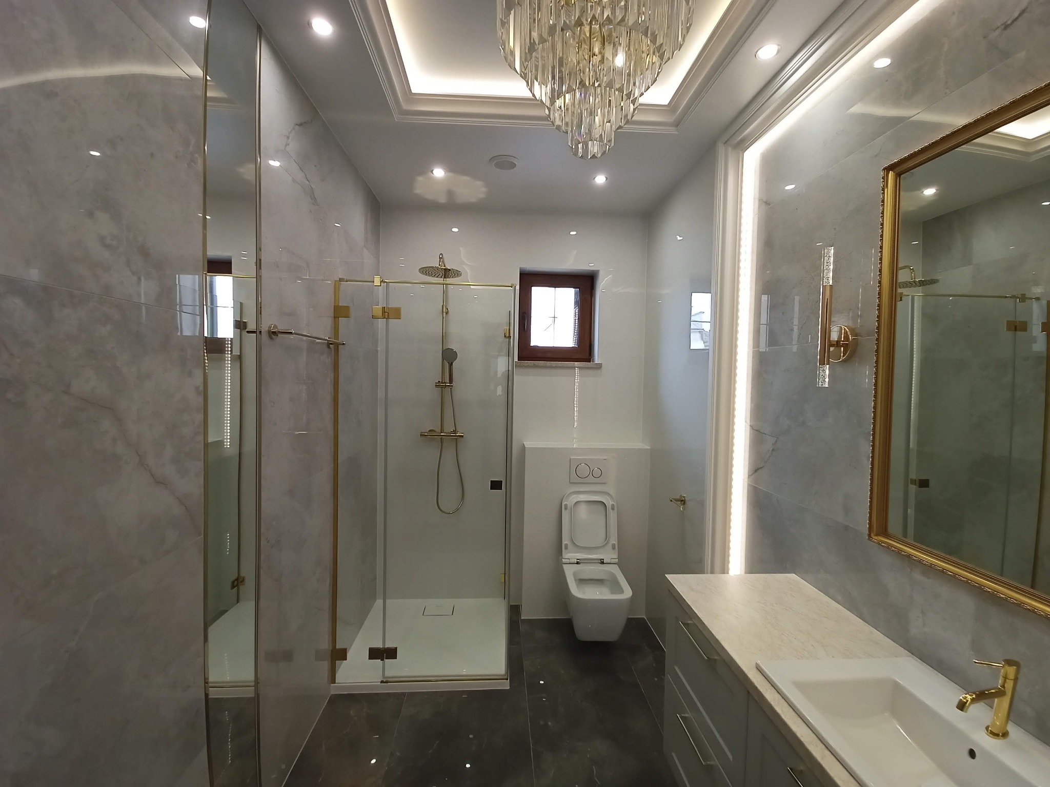 Elegancka łazienka z marmurowymi płytkami, złotymi akcentami w kabinie prysznicowej i armaturze, oraz kryształowym żyrandolem.