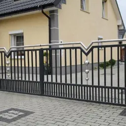 Nowoczesna, czarna brama wjazdowa z dekoracyjnymi, metalowymi kulami, osadzona na kamiennych słupkach. W tle widoczny dom z żółtą elewacją i grafitowym dachem.