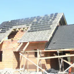 Wznoszony dom z czerwonej cegły z częściowo położonym dachem z czarnej dachówki. Widoczne drewniane elementy konstrukcyjne i rusztowania. Błękitne niebo w tle.