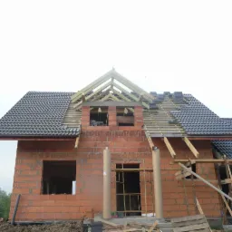 Budowa domu w trakcie: częściowo pokryty dach ciemną dachówką, ściany z czerwonej cegły, widoczne drewniane elementy konstrukcyjne. Budowa w toku.