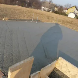 Świeżo wylany, gładki fundament pod budynek z wystającymi prętami zbrojeniowymi i drewnianym szalunkiem, na tle pagórkowatego terenu i zabudowań mieszkalnych.