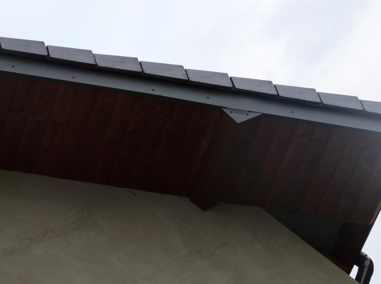 Wykończony okap dachu z drewnianą podbitką i ciemną dachówką, widoczny fragment ściany budynku. Ujęcie od dołu, pochmurne niebo w tle.