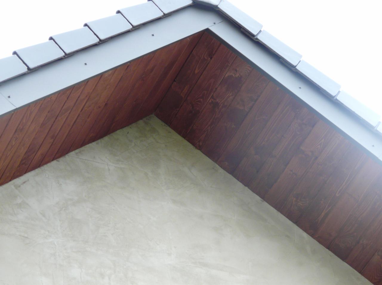 Fragment dachu z brązową drewnianą podbitką i szarymi dachówkami, widoczne mocowania. Tynk na ścianie ma marmurkowy wzór.