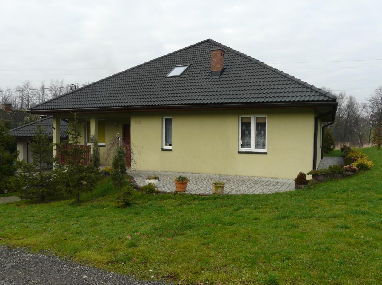 Parterowy dom z ciemną dachówką i ceglanym kominem. Elewacja w odcieniach żółci, małe okna. Uwagę zwraca numer 211 nad drzwiami.