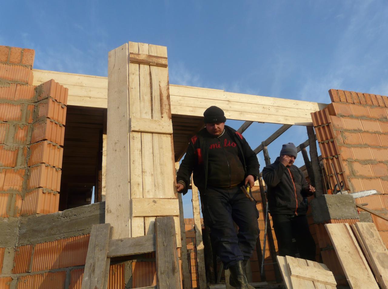 Wznoszenie ścian z czerwonej cegły na budowie domu jednorodzinnego. Widoczni dwaj pracownicy w trakcie pracy i drewniane elementy konstrukcyjne.