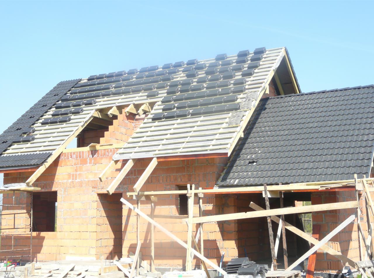 Wznoszony dom z czerwonej cegły z częściowo położonym dachem z czarnej dachówki. Widoczne drewniane elementy konstrukcyjne i rusztowania. Błękitne niebo w tle.