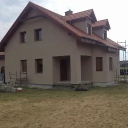 Elewacja domu jednorodzinnego w Kłodawie, świeżo po tynkowaniu, z widocznymi rusztowaniami. Budynek z brązową stolarką okienną i dachem z czerwonej dachówki.