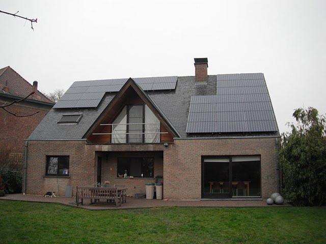 Dom jednorodzinny z panelami słonecznymi na dachu, widok z ogrodu. Na pierwszym planie trawnik, w tle ceglana elewacja i szary dach.