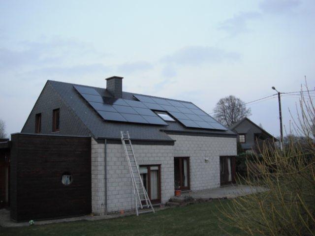 Dom z szarej cegły z panelami słonecznymi na dachu pokrytym łupkiem. Aluminiowa drabina oparta o ścianę. W tle widoczny drugi dom i pochmurne niebo.