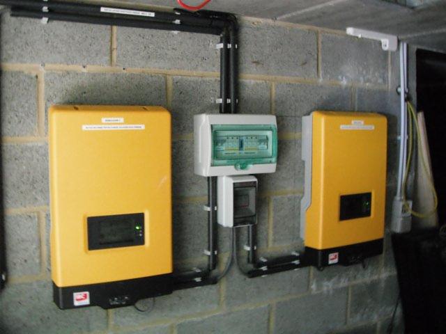 Żółte falowniki i skrzynka elektryczna zamontowane na szarej, betonowej ścianie. Instalacja przewodów w rurkach. Widoczne złącza i elementy mocujące.