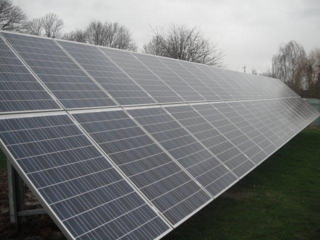 Instalacja paneli słonecznych na trawiastym terenie, widok z boku. Szare niebo w tle, częściowo widoczne drzewa bez liści. Układ paneli ustawiony pod kątem.