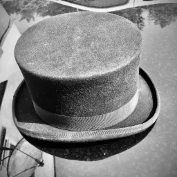 Czarno-białe zdjęcie cylindra z delikatną wstążką, leżącego na błyszczącej, odblaskowej powierzchni, odbijającej budynek i drzewa.