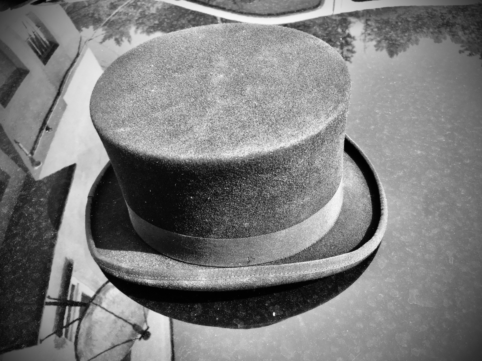 Czarno-białe zdjęcie cylindra z delikatną wstążką, leżącego na błyszczącej, odblaskowej powierzchni, odbijającej budynek i drzewa.