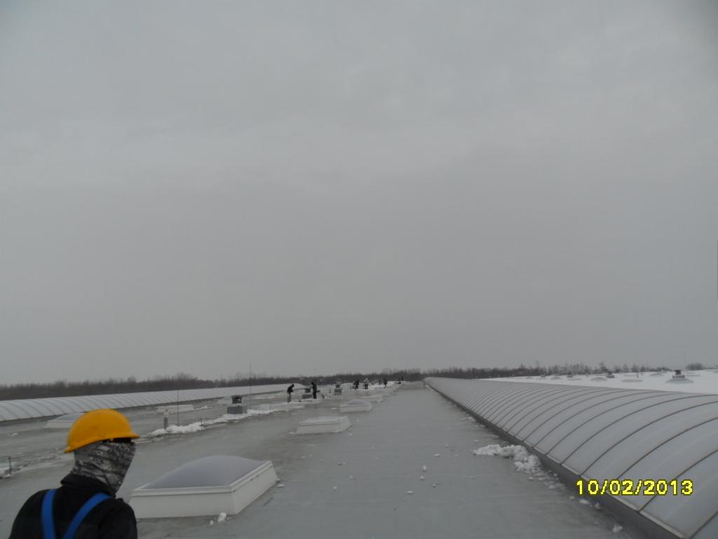 Pracownicy w kaskach na płaskim dachu budynku z świetlikami dachowymi. Widoczny śnieg i pochmurne niebo. Prace na wysokościach w lutym 2013.