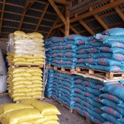 Magazyn z zapasem worków z opałem, ułożonych na paletach, z widoczną drewnianą konstrukcją dachu i metalowymi silosami w tle.