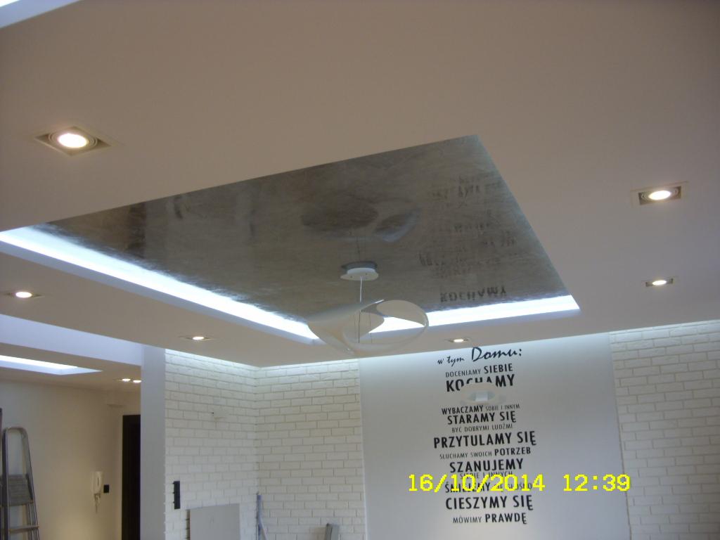 Nowoczesne oświetlenie sufitowe LED z podświetlanym, dekoracyjnym panelem centralnym i punktowymi lampami w białym pomieszczeniu z ceglaną ścianą i sentencjami.